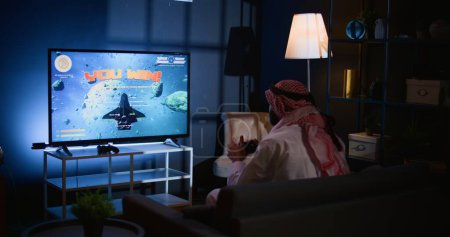 Middle Eastern gamer plays intense classic arcade space shooter videogame. Man relaxing at home using high tech gaming system handheld controller to solve missions in singleplayer game