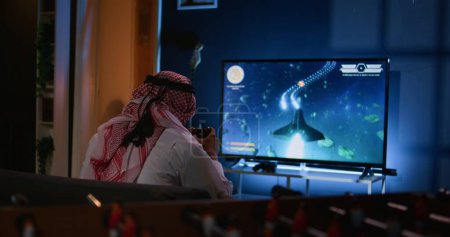 Muslim gamer plays intense classic arcade space shooter videogame, shooting asteroid using laser beams. Arabic man relaxing at home using high tech gaming system to solve missions in singleplayer game