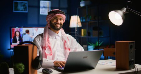 Photo for Happy arabic teleworker answering job emails in stylish apartment. Cheerful muslim employee remotely working, typing data on laptop with opened tv as background noise, handheld camera shot - Royalty Free Image