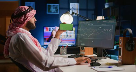 Photo for Arab man watching video on green screen cellphone while looking at business annual report charts on computer. Worker spending time on chroma key mobile phone while solving job tasks at home - Royalty Free Image