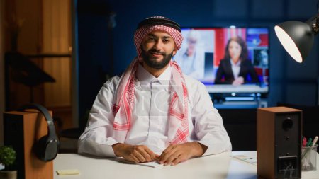 Photo for Cheerful Arab man paying attention in elearning seminar teleconference with teacher. Middle Eastern student in online videocall greeting tutor, listening to lesson while writing down homework tasks - Royalty Free Image