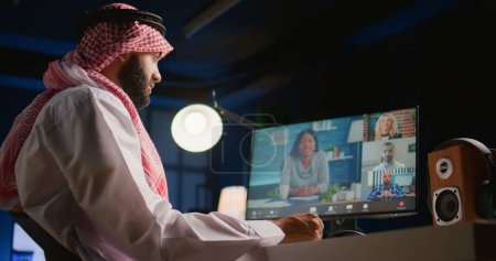 Téléchargez les photos : Superviseur arabe tenant une téléconférence en ligne avec des sous-alternes de travail à distance, vérifiant l'avancement des tâches. Dirigeant d'entreprise musulman à la maison discutant avec les employés pendant le vidéocall - en image libre de droit