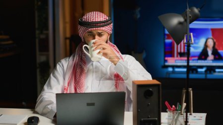 Téléchargez les photos : Candidat arabe profitant d'une boisson tout en étant interviewé à distance par l'équipe des ressources humaines. Musulman boit du café tout en obtenant ses pouvoirs vérifiés lors de la réunion de vidéoconférence en ligne - en image libre de droit