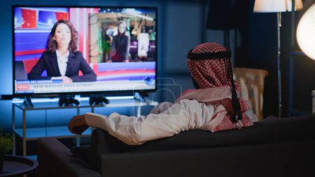 Der arabische Mann lümmelte auf dem gemütlichen Sofa und sah Nachrichten, während er sich nach einem anstrengenden Arbeitstag zu Hause entspannte. Muslim chillt auf Couch und schaut auf TV-Sender, der aktuelle Ereignisse zeigt