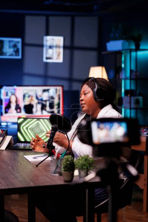 Foto de Mujer negro crea vlogs utilizando cámara profesional mientras que la producción de podcasts con equipo de audio. Periodista mujer entrevista a invitados en su estudio, filmando con un dispositivo en un trípode. - Imagen libre de derechos