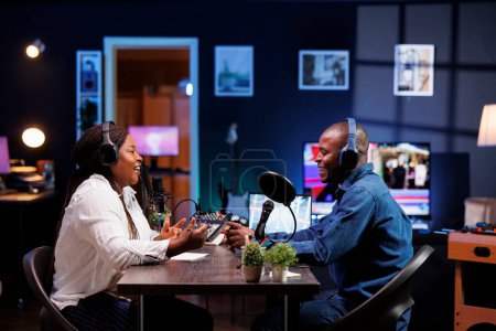 Photo for Cheerful african american couple having a conversation at home, and using audio equipment for podcast episode. Joyful male and female influencers wearing wireless headphones and talking into mics. - Royalty Free Image