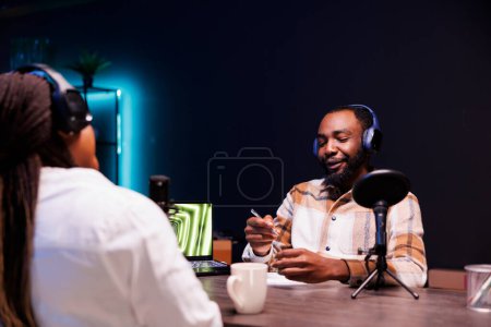Photo for African american couple wearing wireless headphones are making a podcast at home using audio gadgets. Black male host interviewing female blogger for his talk show online audience. - Royalty Free Image