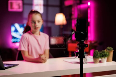 Focus on cellphone on tripod used by kid shooting product unboxing for streaming channel on online platforms. Child in blurry background warmly welcoming fans, filming reviews with mobile phone
