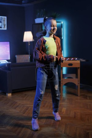 Young girl dancing in dimly lit home studio interior, producing content for online channel. Generation Z youngster doing viral dance choreography in apartment illuminated by RGB lights