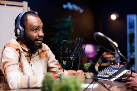 Photo for Man talk show host using audio equipment and having a friendly interview with an upcoming artist at home studio. African american content creator with headphones and microphone is creating a podcast. - Royalty Free Image