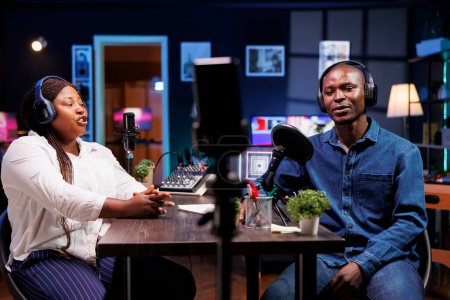 African American content creator couple talks tech in their home studio, entertaining a wide online audience with engaging discussions. Black vloggers using mobile device for recording radio program.