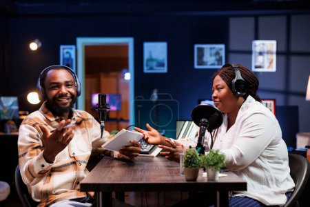Ein bärtiger Mann lacht während eines Comedy-Podcasts, während die Moderatorin mit einer afroamerikanischen Bloggerin scherzt. Männlicher Journalist unterhält sowohl Gäste als auch Zuschauer, die von zu Hause aus zusehen.
