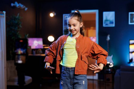 Young gen Z content creator in dimly lit home studio dances for online followers. Small girl does viral dance choreography in neon lit living room, filming with cellphone
