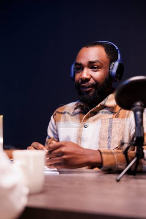 Photo for African american man using audio equipment to create an online radio show at home. Young black male content creator with wireless headphones and speaking into a microphone, is making a podcast. - Royalty Free Image