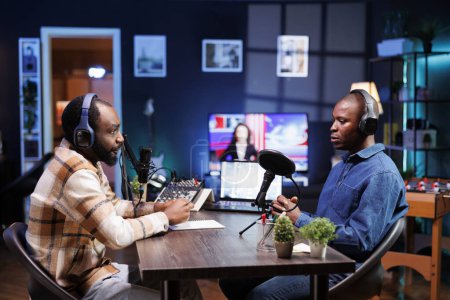 Photo for Sitting at table using the audio equipment are two african american men discussing and addressing their online audience. Interviewer asking male influencer questions, recording a radio show at home. - Royalty Free Image