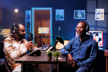 Program host and guest conversing in an elaborate home studio with neon lighting. African american influencers use professional equipment to broadcast a discussion on online podcast episode.