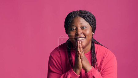 Atemberaubende schwarze Frau blickt in die Kamera und fleht wirklich mit ihren Händen zusammen. Junge Afroamerikanerin mit ansprechender und fordernder Körpersprache vor rosa Hintergrund.
