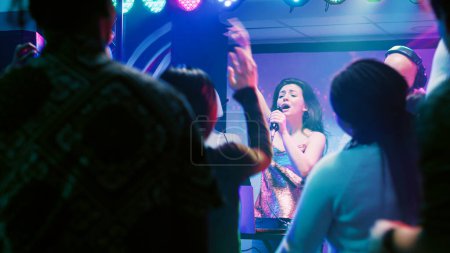 Confident woman doing karaoke on stage, singing songs with male DJ in front of people at nightclub. Happy girl having fun with live perfromance next to audio mixing station in club.