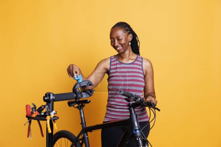 Energetische schwarze Frau, die Fahrradeinstellungen mit Werkzeug vornimmt und Komponenten inspiziert. Sportbegeisterte Afroamerikanerin untersucht Reparaturständer für kaputte Fahrradrahmen. Isolierter gelber Hintergrund.
