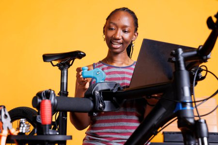 Junge schwarze Frau inspiziert und repariert beschädigtes Fahrrad gründlich mit Anweisungen vom Laptop. Gesunde Afroamerikanerin recherchiert im Internet für die Wartung von Fahrrädern.