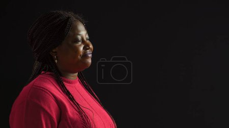 Photo for Positioned against isolated black backdrop young woman wears downcast facial expression. Side-view portrait of african american fashion blogger in a mournful stance against blank background. - Royalty Free Image