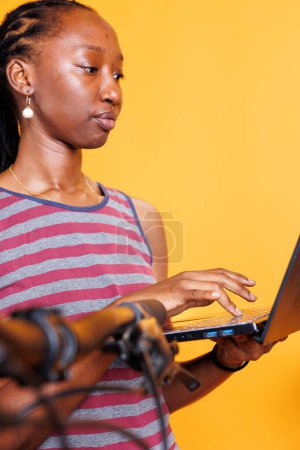 Photo for Dedicated young lady engrossed in inspecting and servicing bicycle with laptop. African American woman resolves broken bike with digital pc, ensuring safety and functionality. Close up shot. - Royalty Free Image