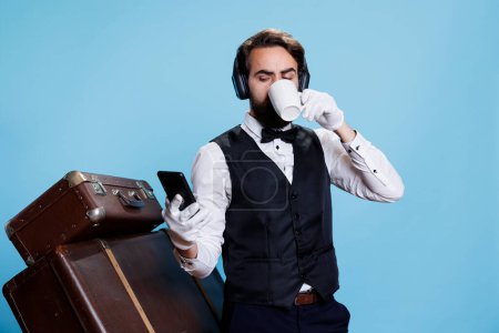 Hotel porter drinks coffee and texting, reading messages on smartphone and listening to music on headphones. Doorkeeper bellboy with headset enjoying drink and browsing webpages.