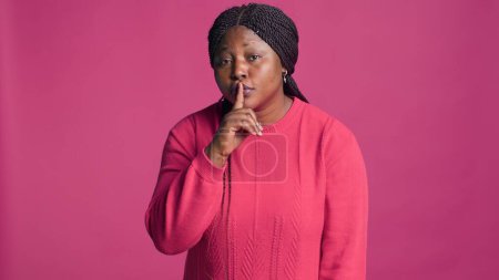 African american woman standing in front of isolated background expressing with hand gestures. Female fashion blogger looking at camera and saying quiet shush with finger on her mouth.