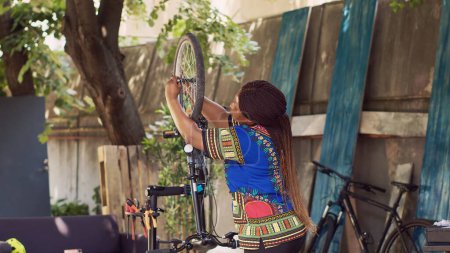 Photo for Active african american woman cautiously loosening and removing damaged bicycle wheel. Sports-loving female cyclist carefully dismantling tire from bike front fork for maintenance in home yard. - Royalty Free Image