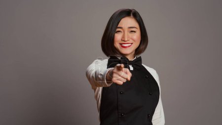 Photo for Asian waitress pointing at camera to choose you over grey background, wearing uniform with bow and apron. Woman catering worker choosing someone to work at a restaurant. Camera B. - Royalty Free Image