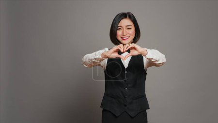 Photo for Hotel administrator shows heart shape symbol on camera, expressing romantic gesture and being flirty. Asian receptionist presenting true honest feelings, does romance sign in studio. Camera A. - Royalty Free Image