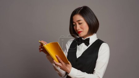 Photo for Asian receptionist reading a literature book over grey background, enjoying lecture hobby with a novel story in studio. Hotel concierge employee reads a fiction tale for knowledge. Camera B. - Royalty Free Image