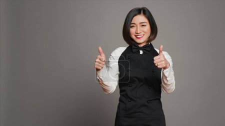 Smiling waitress giving thumbs up sign in front of camera, working in a five star restaurant to serve tables. Asian server with apron showing approval with okay symbol in studio, agreement. Camera A.