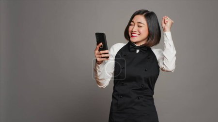 Asian server feeling happy after reading good news on phone, having a lot of clients with reservations for dinner. Waitress with apron using smartphone to see table bookings, enjoys work. Camera A.