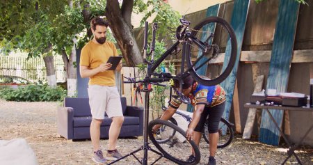 Caucasian man standing with smart device while african american woman dismantles damaged bike wheel. Couple using digital tablet for researching of bicycle adjustments for summer maintenance.