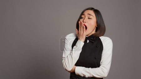 Photo for Exhausted waitress with apron yawning over grey background, feeling overworked and dealing with burnout after multiple restaurant events. Asian catering worker feeling tired in studio. Camera B. - Royalty Free Image