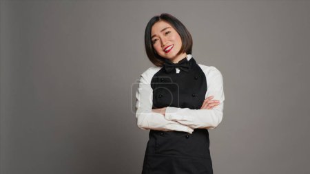 Photo for Asian restaurant employee serving clients at tables, offering luxury catering services with tray on camera. Waitress working in gourmet industry, wearing apron and bow in studio. Camera A. - Royalty Free Image