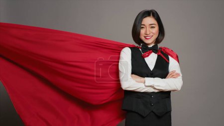 Téléchargez les photos : La réceptionniste asiatique posant avec une cape de super-héros rouge à la caméra, se sentant confiante et puissante alors qu'elle porte un manteau en héros. Concierge de l'hôtel debout les bras croisés. Caméra A. - en image libre de droit