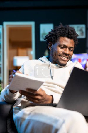 Unternehmer, der von einer neonbeleuchteten Wohnung aus arbeitet und Firmenideen aufschreibt, während er sich Videos auf seinem Laptop ansieht. BIPOC-Mann freut sich über Startup-Vorschlag und notiert Plan mit Stift