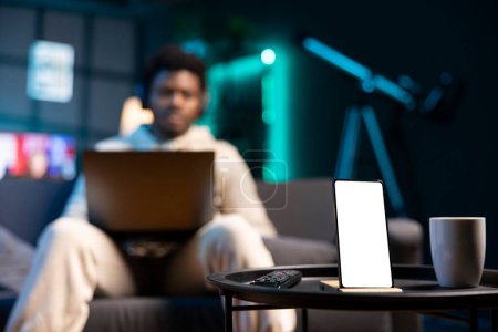 Photo for Focus on mockup device with freelancer in blurry background working on laptop and listening music. Close up of isolated screen cellphone in front of teleworker wearing headphones, doing tasks - Royalty Free Image