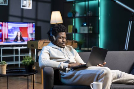 Foto de Hombre afroamericano sentirse aburrido, desplazarse por Internet utilizando el ordenador portátil mientras escucha música, relajarse durante el fin de semana. Persona con auriculares, navegando por la red en casa - Imagen libre de derechos