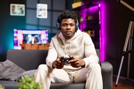 Man sitting on couch at home, relaxing by playing videogames on gaming console. BIPOC gamer unwinding in living room by competing in online multiplayer esports tournament