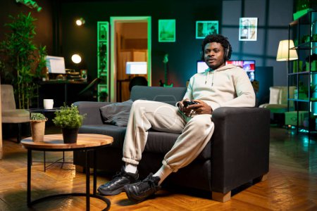 Afroamerikanischer Mann in schummriger Wohnung, der Videospiele spielt, sich entspannt und Spaß hat. Gamer kämpft in der Freizeit zu Hause gegen Feinde im Multiplayer-Spiel auf Spielkonsole