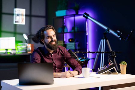Happy internet star using quality mic in studio to discussing with viewers. Smiling man sitting at table in front of laptop using high tech recording gear to record social media video