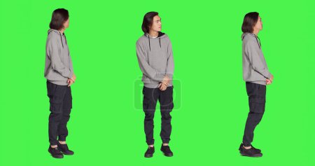 Young person looks at wristwatch standing against full body greenscreen backdrop, acting impatient on camera. Asian guy feeling stressed and irritated, waiting for a certain thing.