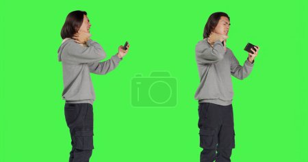 Photo for Asian man filming video on phone, wearing cervical neck brace over greenscreen in studio. Young adult recording montages or shots while he deals with bone fracture using foam collar brace. - Royalty Free Image