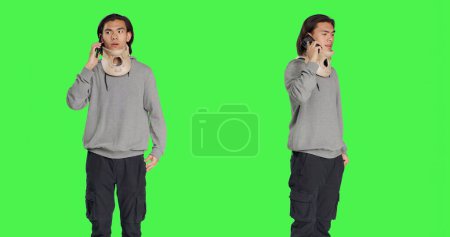 Photo for Young adult with neck collar answers call on smartphone, talking to people about his bone fracture accident. Hurt person wearing cervical foam brace discussing on telephone line, greenscreen. - Royalty Free Image