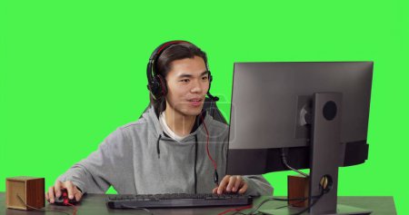 Young person sitting at desktop office, having fun with friends while enjoying online multiplayer games with headphones. Over greenscreen, asian guy competes in role playing contest.