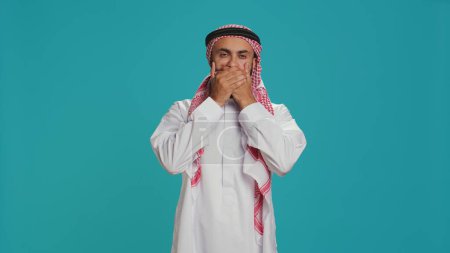 Muslim adult covers eyes, ears and mouth in studio, showcasing three wise monkeys metaphor sign. Middle eastern guy presenting dont hear, see or speak concept symbol in arabic clothes.