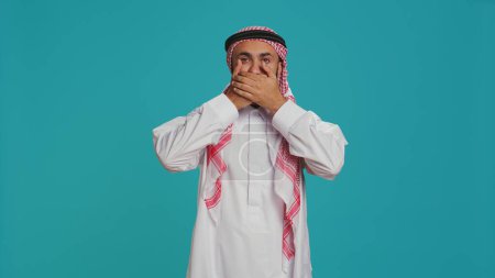 Photo for Male model covers eyes, ears and mouth on camera, presenting three wise monkeys symbol. Middle eastern person creating dont hear, see or speak metaphor sign, islamic clothing. - Royalty Free Image